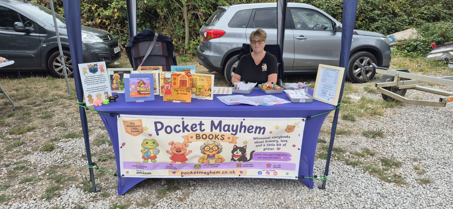 Carol McMahon seated behind her Pocket Mayhem book stall at an outdoor market, with the Pocket Mayhem banner and a display of children's books visible